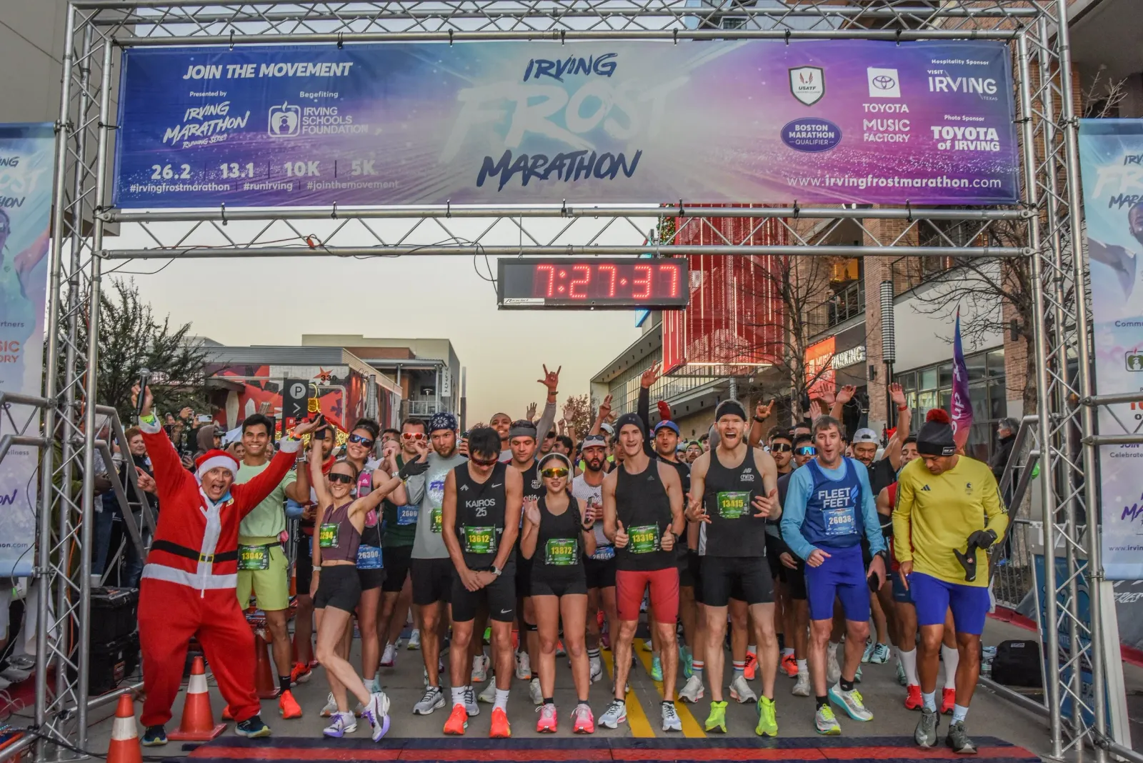 A group of runners, some in festive attire including a Santa suit, cheer at the start line of the Irving Frost Marathon under a large banner and a digital clock.
