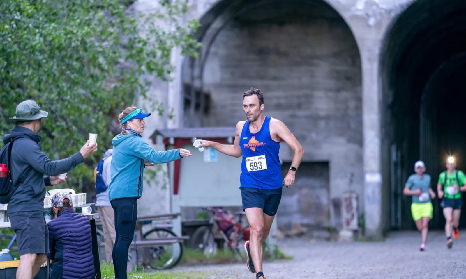 A male runner in a blue tank top with bib number 593 receives a cup from a volunteer at an aid station, with other runners in the background near tunnel entrances.