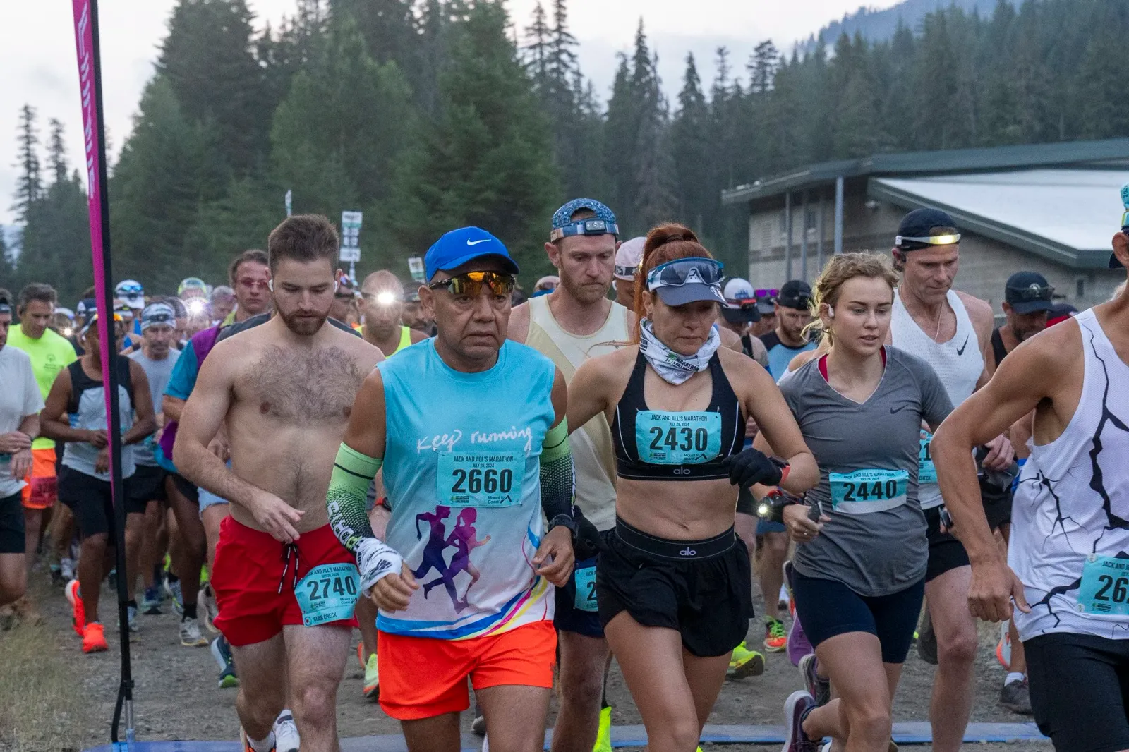 A group of runners at the start line of the Jack and Jill's Marathon, with trees and a building in the background.