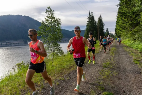 Runners on a gravel path next to a body of water, with trees and hills in the background.