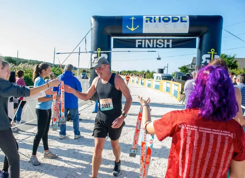 A male runner smiles as he crosses the finish line and receives a medal from volunteers at the Rhode Island Races event.