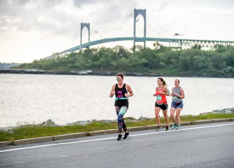 Three female runners on a road course with a large suspension bridge in the background over a body of water.