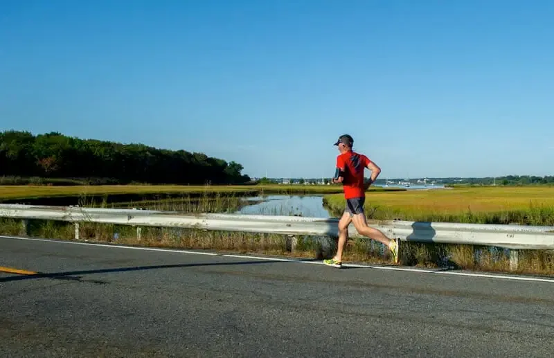 A runner in a red shirt and dark shorts runs along a road next to a marshy waterway with trees and boats in the distance under a clear blue sky.