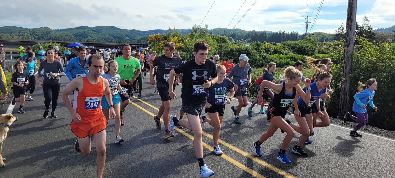 A group of runners, including men, women, and children, are starting a race on a paved road with hills and trees in the background.