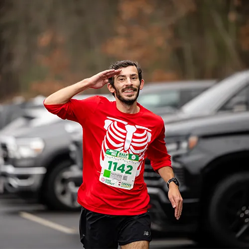 A male runner in a red long-sleeved shirt with a skeleton ribcage design and bib number 1142 salutes while running.