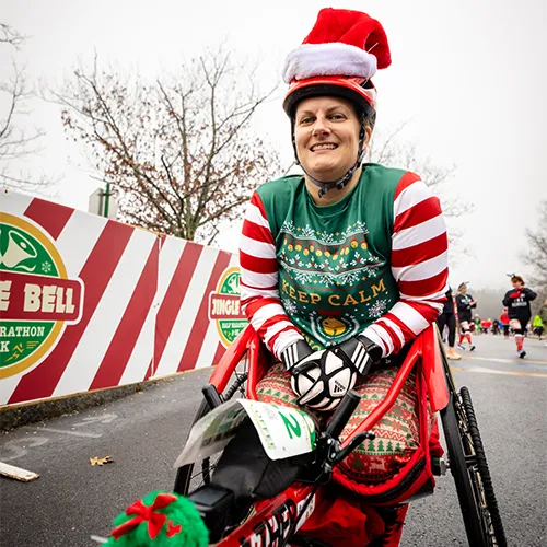 A participant in a wheelchair wearing a Santa hat and a festive holiday-themed running outfit smiles at the camera during the Jingle Bell Half Marathon.