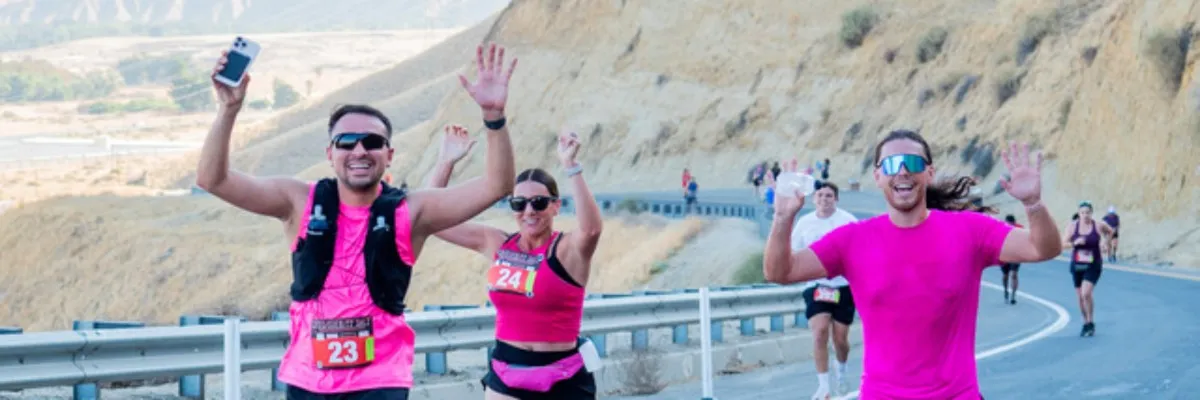 Three runners, two in the foreground with arms raised in celebration and one in the background, run along a paved road with a scenic, hilly landscape.