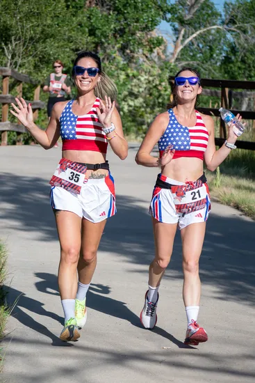 Two female runners in patriotic-themed running outfits smile and wave while running on a paved path.
