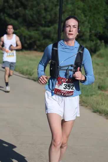A female runner wearing a blue long-sleeved shirt, black vest, and white shorts runs on a paved path, with another runner in the background.