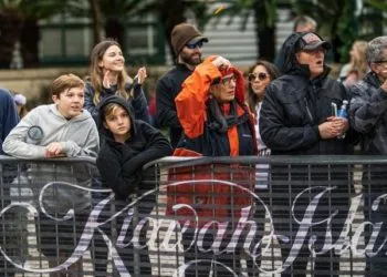 Spectators watch a race from behind a barrier, some looking engaged and others shielding themselves from the elements.
