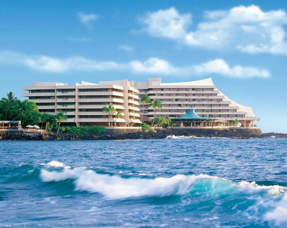 A large, modern hotel building sits on a rocky coastline with palm trees, overlooking the blue ocean with waves in the foreground.