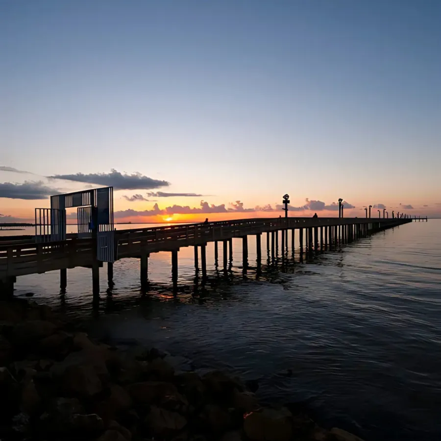 A wooden pier extends into the calm water of a bay at sunrise, with the sun peeking over the horizon and casting a warm glow on the clouds.