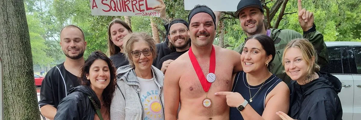 A group of smiling people, one shirtless man wearing a race medal, celebrating together after a race.