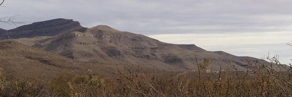 A wide shot of a rugged mountain range under a cloudy sky, with dry brush in the foreground.
