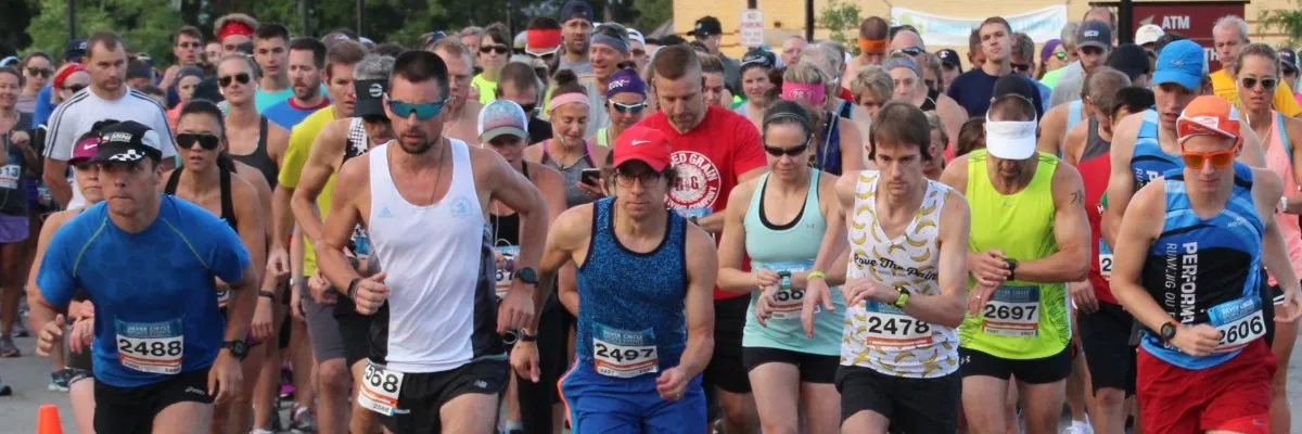 A large group of runners in the foreground and background, lined up at the start of a race, with bib numbers visible on their shirts.