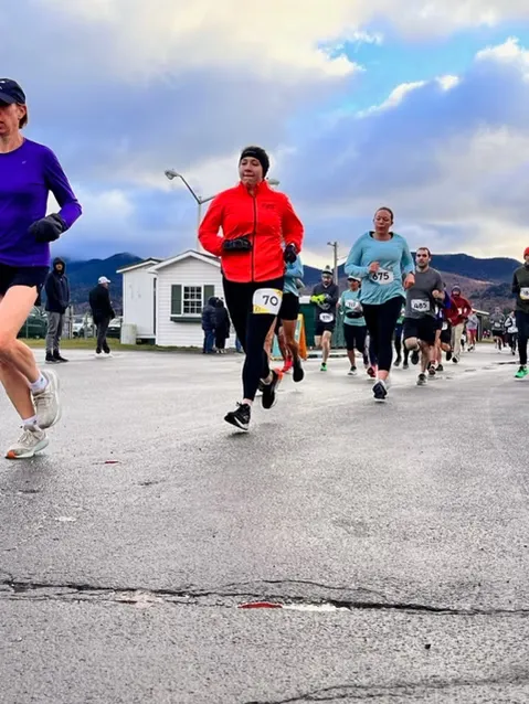 Runners in colorful jackets and athletic wear running on a wet road, with mountains in the background under a cloudy sky.
