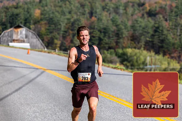 A male runner with bib number 283 runs on a road with a scenic, tree-covered hillside in the background.