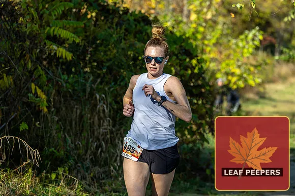 A female runner with her hair in a bun, wearing sunglasses and a white tank top, runs on a tree-lined course.