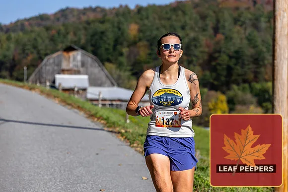 A female runner with sunglasses and a bib number runs on a road with a barn and trees in the background.
