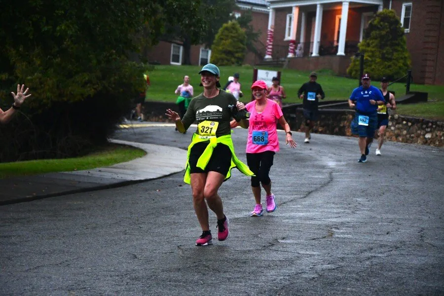 Two female runners smiling and waving as they run on a paved road during a race, with other runners in the background.