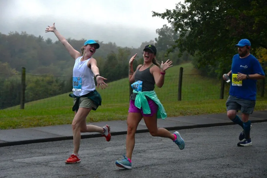 Three runners on a road, two women in the foreground with arms raised in celebration, and a man in the background.