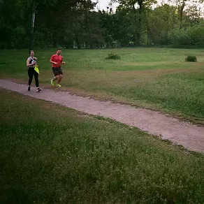 Two runners, a woman and a man, jogging on a dirt path through a grassy park with trees in the background.
