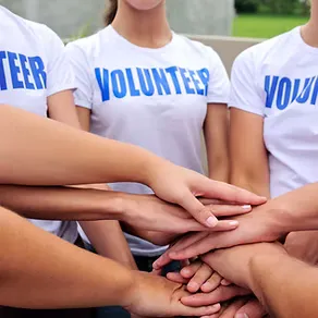 Group of volunteers wearing matching t-shirts with the word "VOLUNTEER" and placing their hands together in a show of unity.