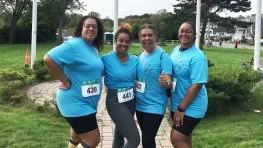 Four women in matching blue t-shirts pose for a photo at a race, smiling and giving thumbs up.
