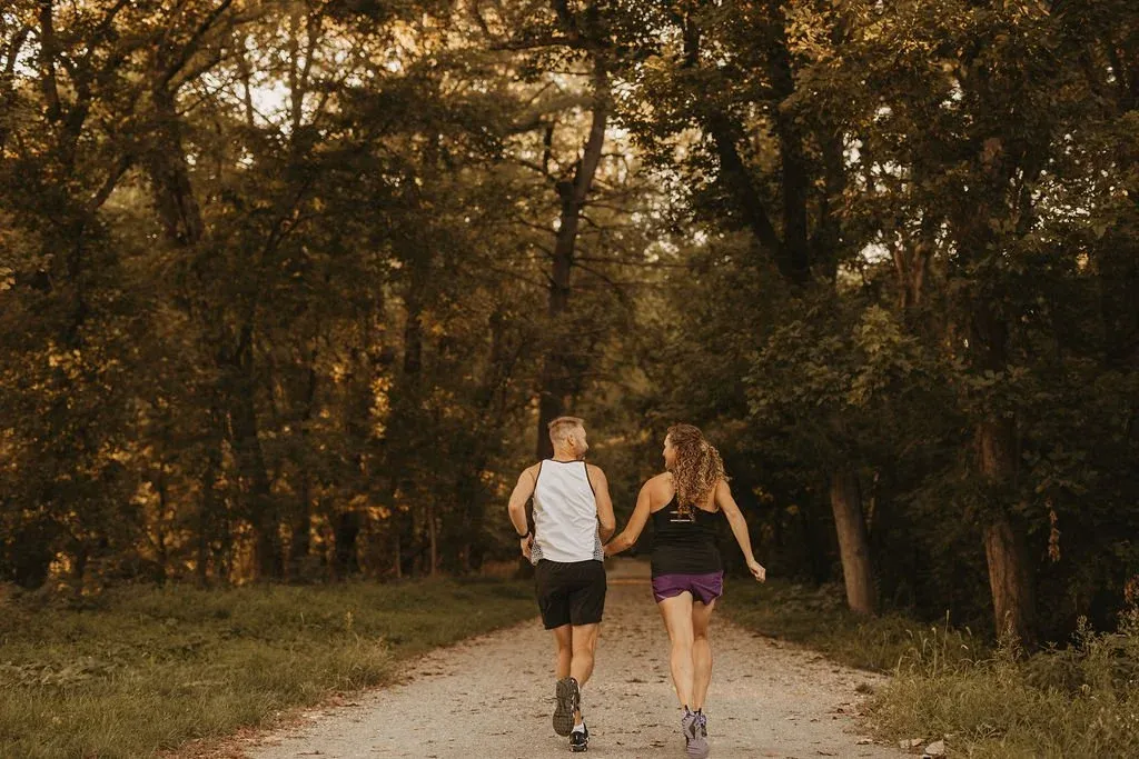 A man and a woman running hand-in-hand on a dirt path through a sun-dappled forest.