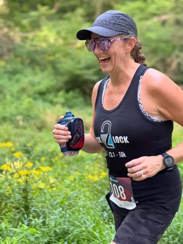A female runner wearing a black tank top with a race logo, a race bib number 108, a baseball cap, and sunglasses, running on a trail with yellow flowers in the background.