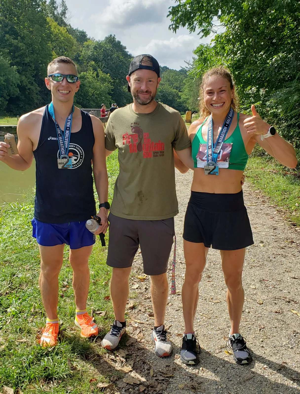 Three smiling runners, two wearing medals, pose for a photo after a race on a gravel path lined with trees.