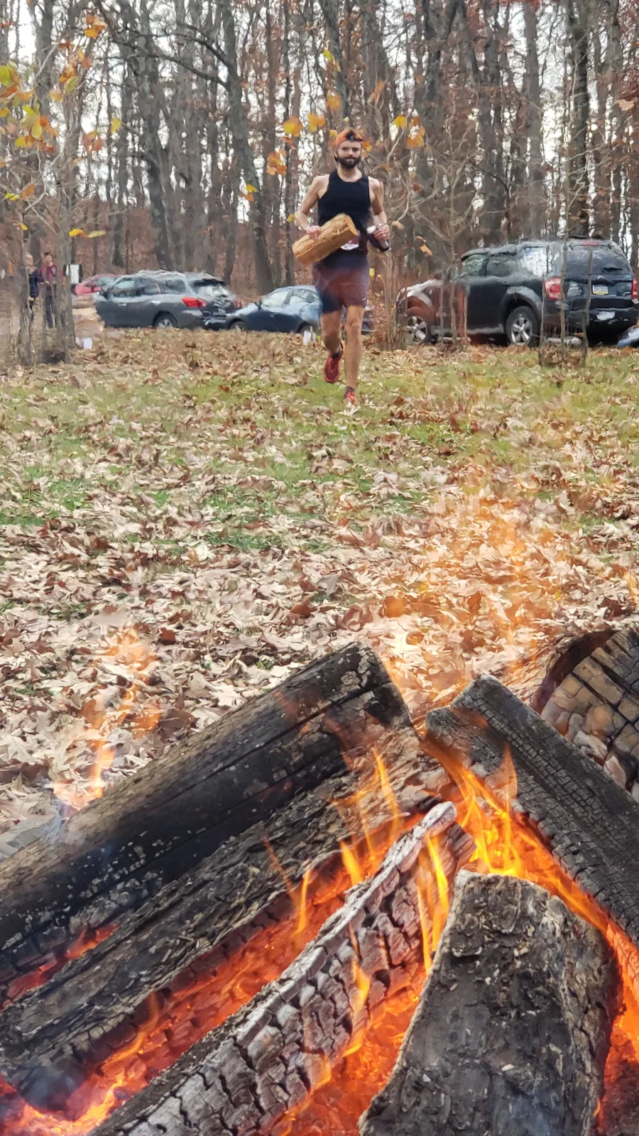 A runner carrying a log runs through a forest with fallen leaves on the ground and a campfire in the foreground.