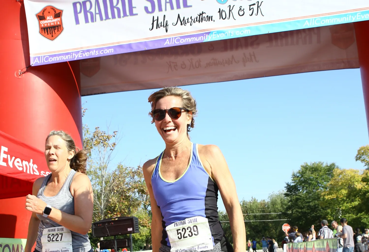A woman laughs joyfully at the start line of the Prairie State Half Marathon, 10K & 5K, with another runner beside her.