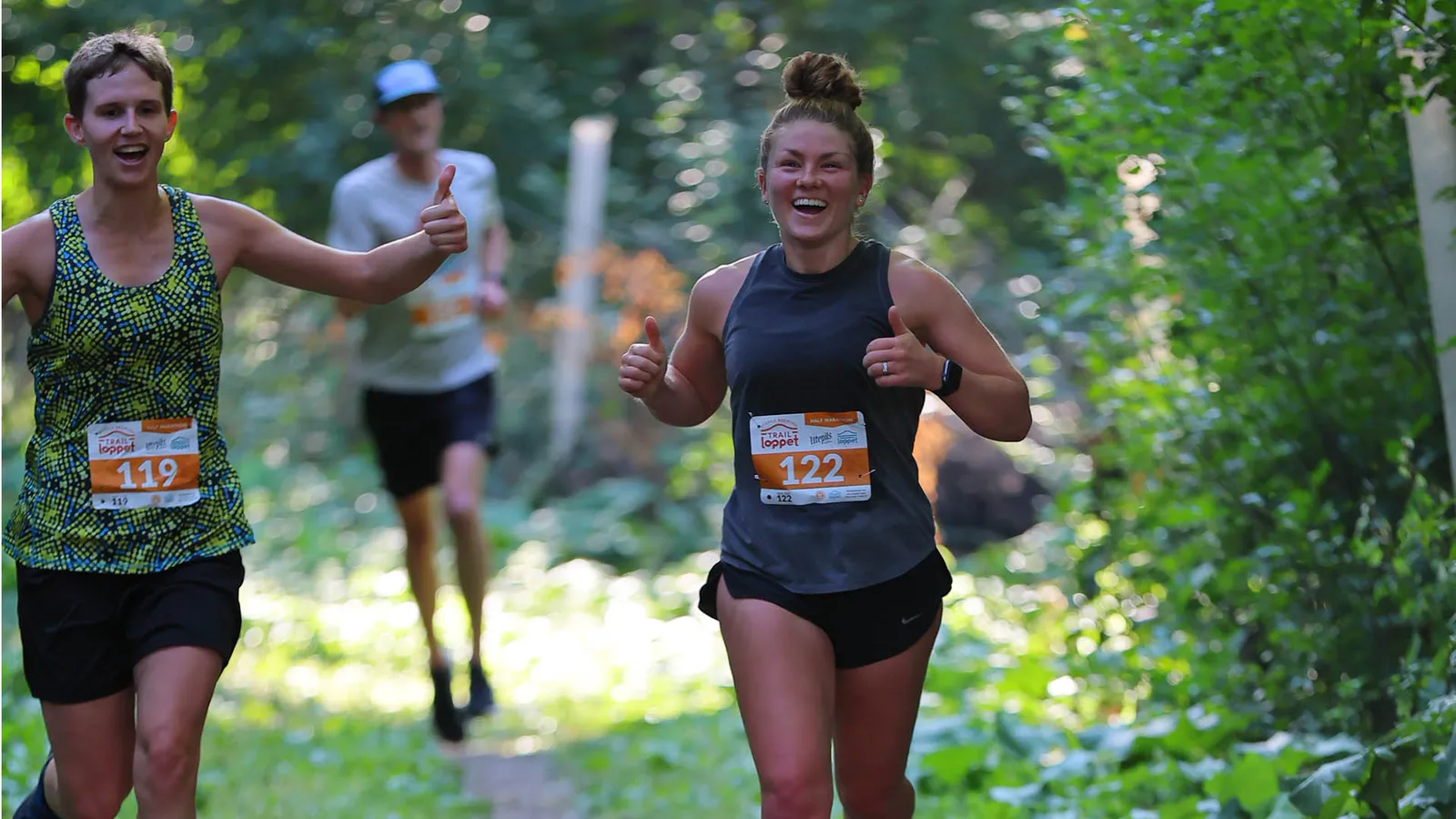 Two female runners smiling and giving thumbs up while running on a wooded trail.