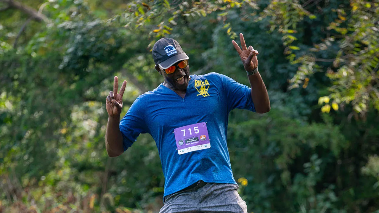 A runner wearing a blue shirt with a bib number 715, sunglasses, and a cap, smiles and holds up peace signs while running on a trail.