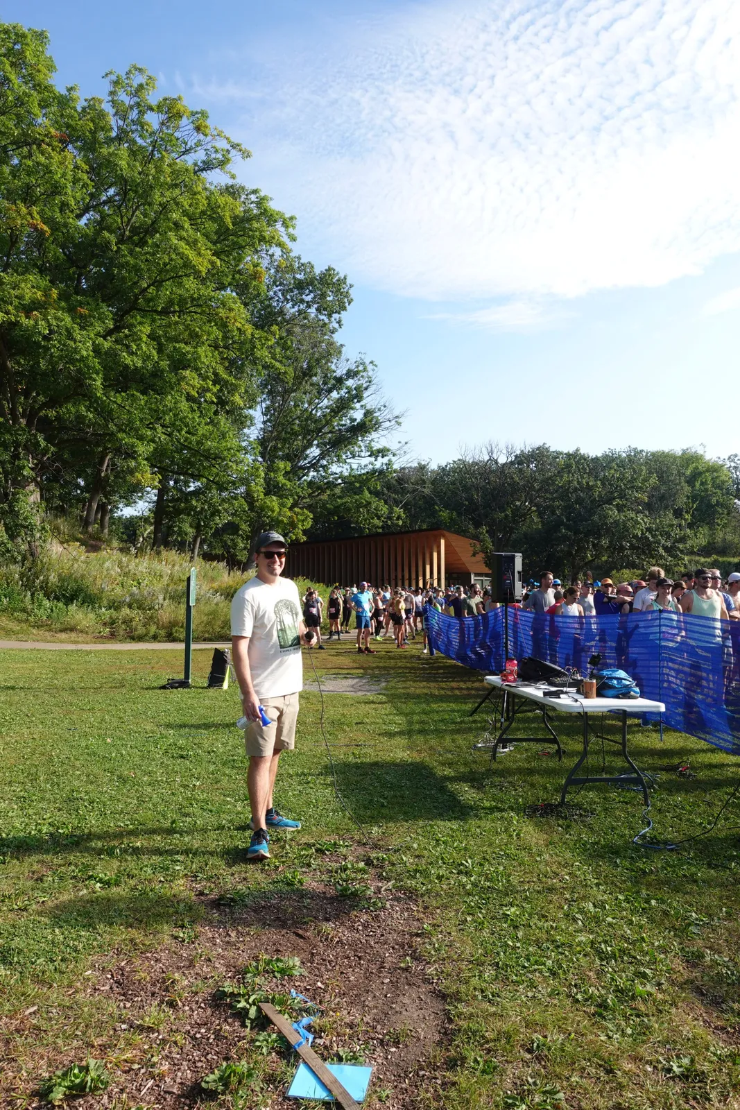 A man with a microphone stands in the foreground, smiling, with a crowd of runners gathered behind a blue barrier in the background.