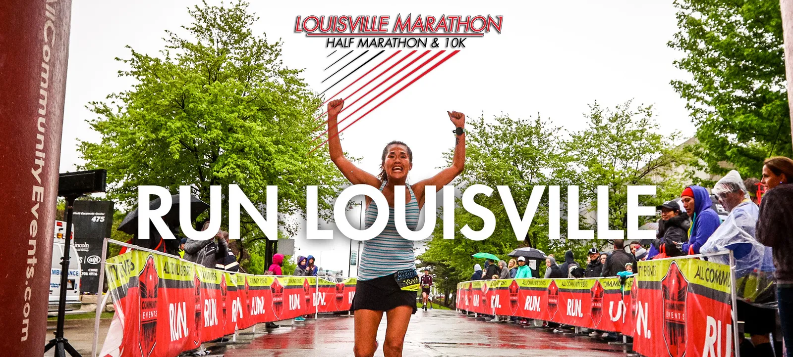 A female runner with arms raised in celebration crosses the finish line of the Louisville Marathon, with spectators watching in the background.