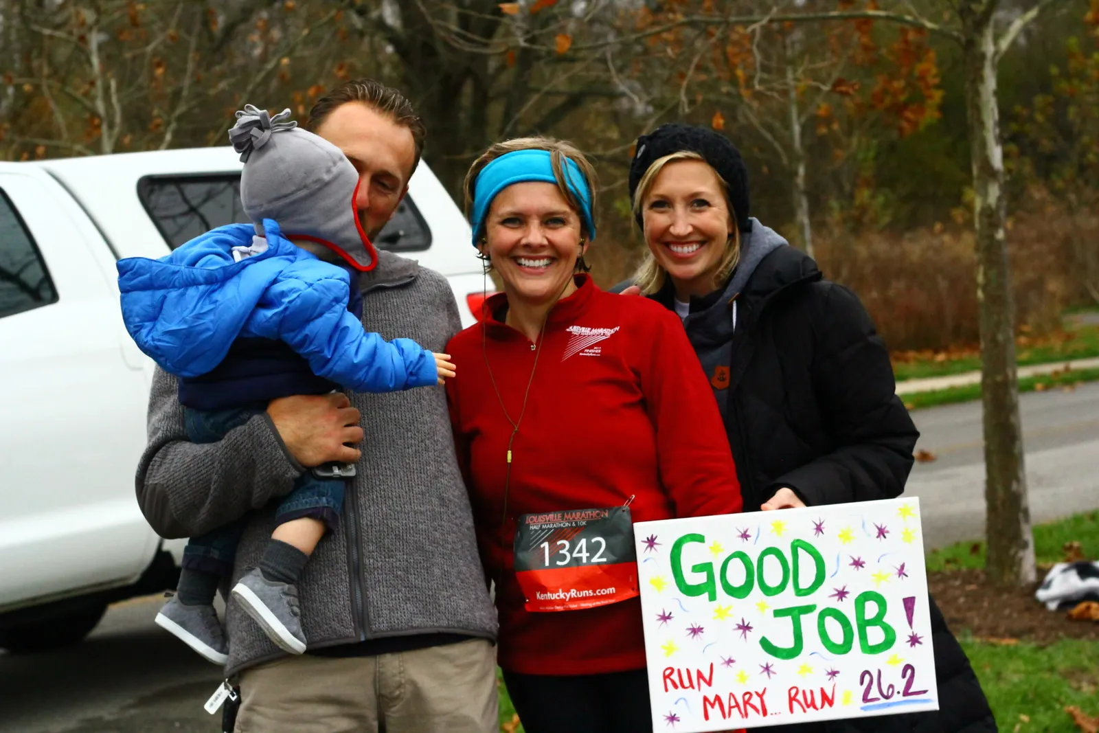 A family smiles while holding a 'Good Job! RUN MARY RUN 26.2' sign at a race event.