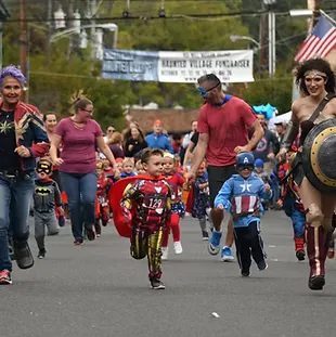 A group of children and adults dressed as superheroes run down a street, with some adults cheering them on.