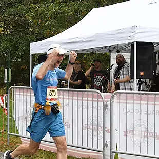 A male runner in a blue shirt and shorts pumps his fist in celebration as he crosses the finish line, with a live band performing in the background.