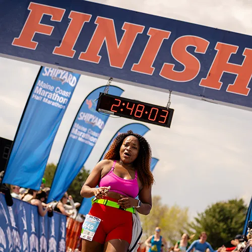 A female runner in a pink top and red shorts crosses the finish line of the Maine Coast Half Marathon, with the race clock showing 2:48:03.