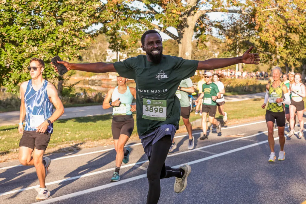 A runner with arms outstretched and a big smile runs towards the camera, with other runners and spectators in the background.