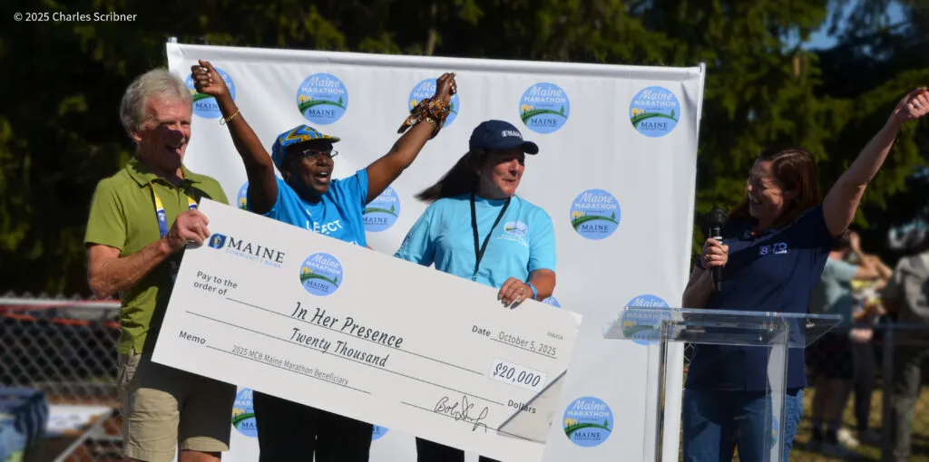 Four people smiling and holding a large ceremonial check for $20,000, with a Maine Marathon banner in the background.