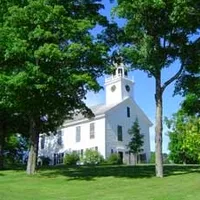 White church with a steeple surrounded by green trees and grass under a blue sky.