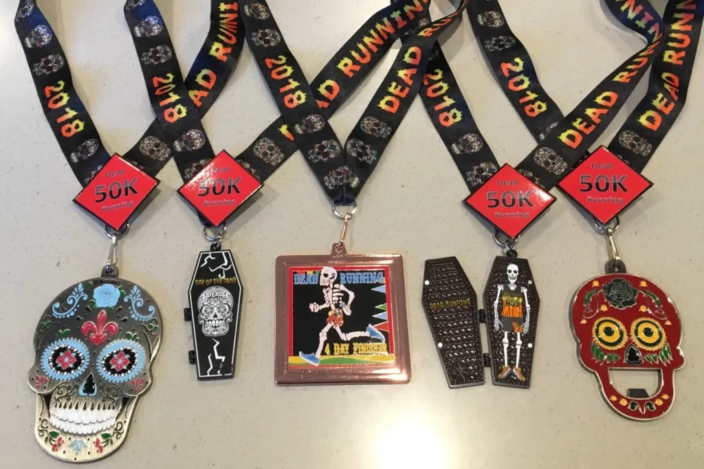 A collection of unique race medals, including skull-shaped and coffin-shaped designs, with ribbons that say "Dead Running 2018 50K".