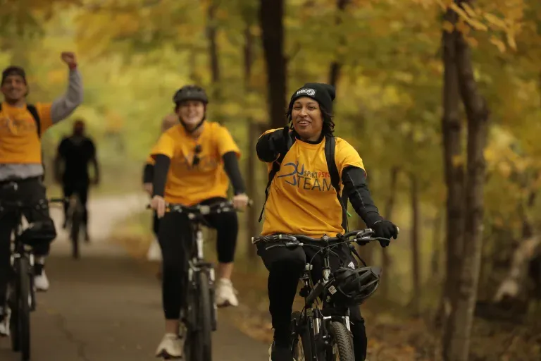 A group of people smiling and riding bicycles on a paved path surrounded by trees with autumn foliage.