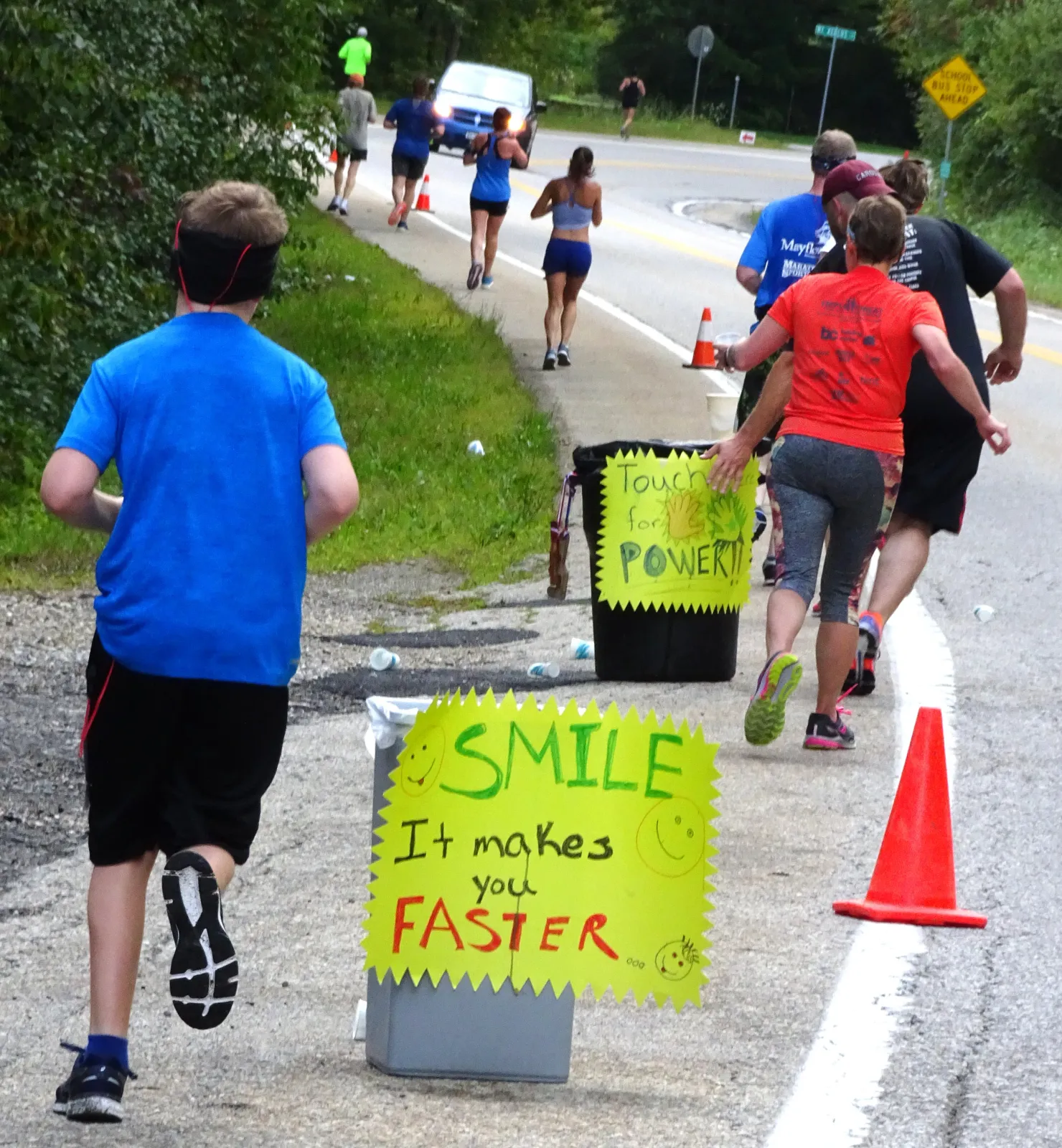Runners on a road course with a sign that says 'SMILE It makes you FASTER'.