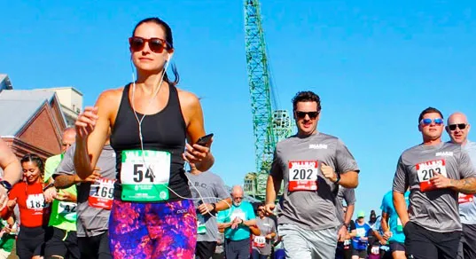 Runners in a race, with a woman in the foreground wearing sunglasses and headphones, and men behind her wearing grey shirts with bib numbers.