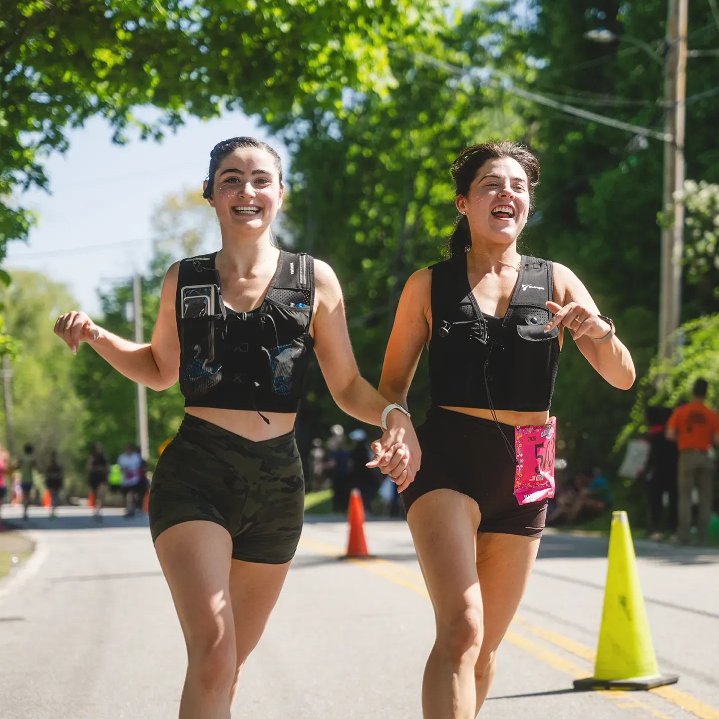 Two female runners smiling and holding hands while running on a road course, with trees and spectators in the background.