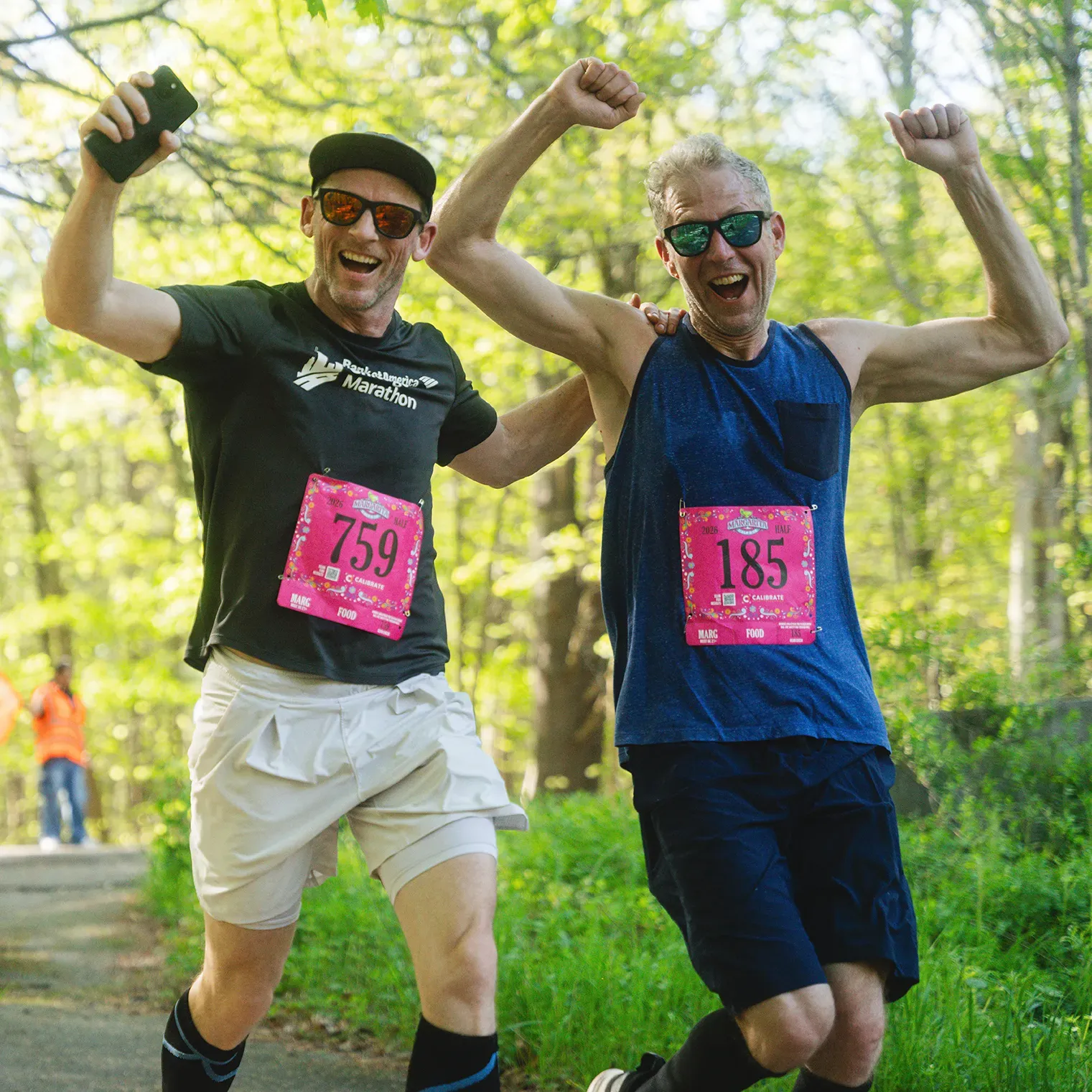 Two male runners, one holding up a phone and the other with arms raised in celebration, run on a wooded path during a race, both wearing bib numbers and smiling.
